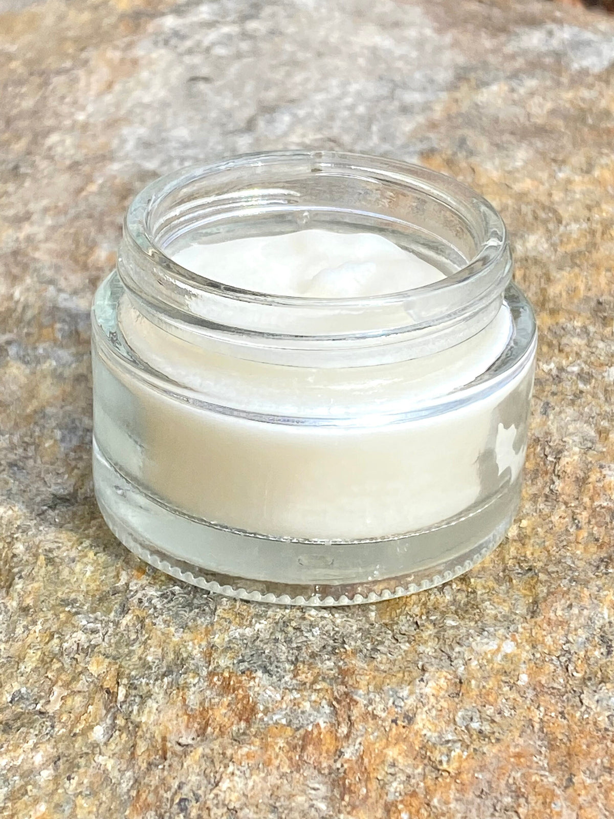 InspireByMK natural all-purpose balm in open clear glass container showing smooth creamy consistency, pure organic skincare product for face, lips, and body on textured stone background