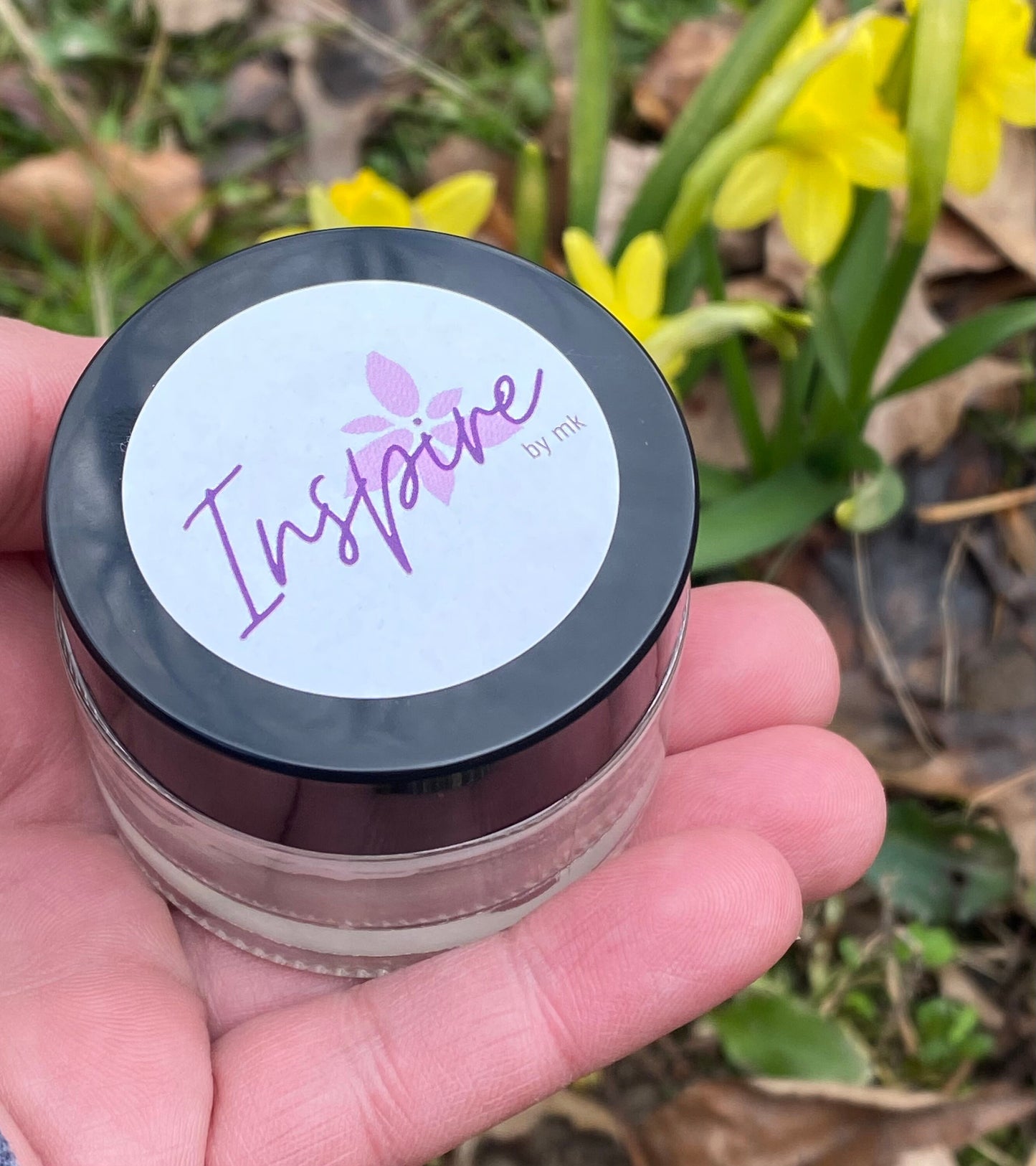 InspireByMK natural balm held in hand showing small glass jar with black screw-on lid and purple Inspire branding label, outdoor garden setting with yellow daffodils, portable skincare balm product