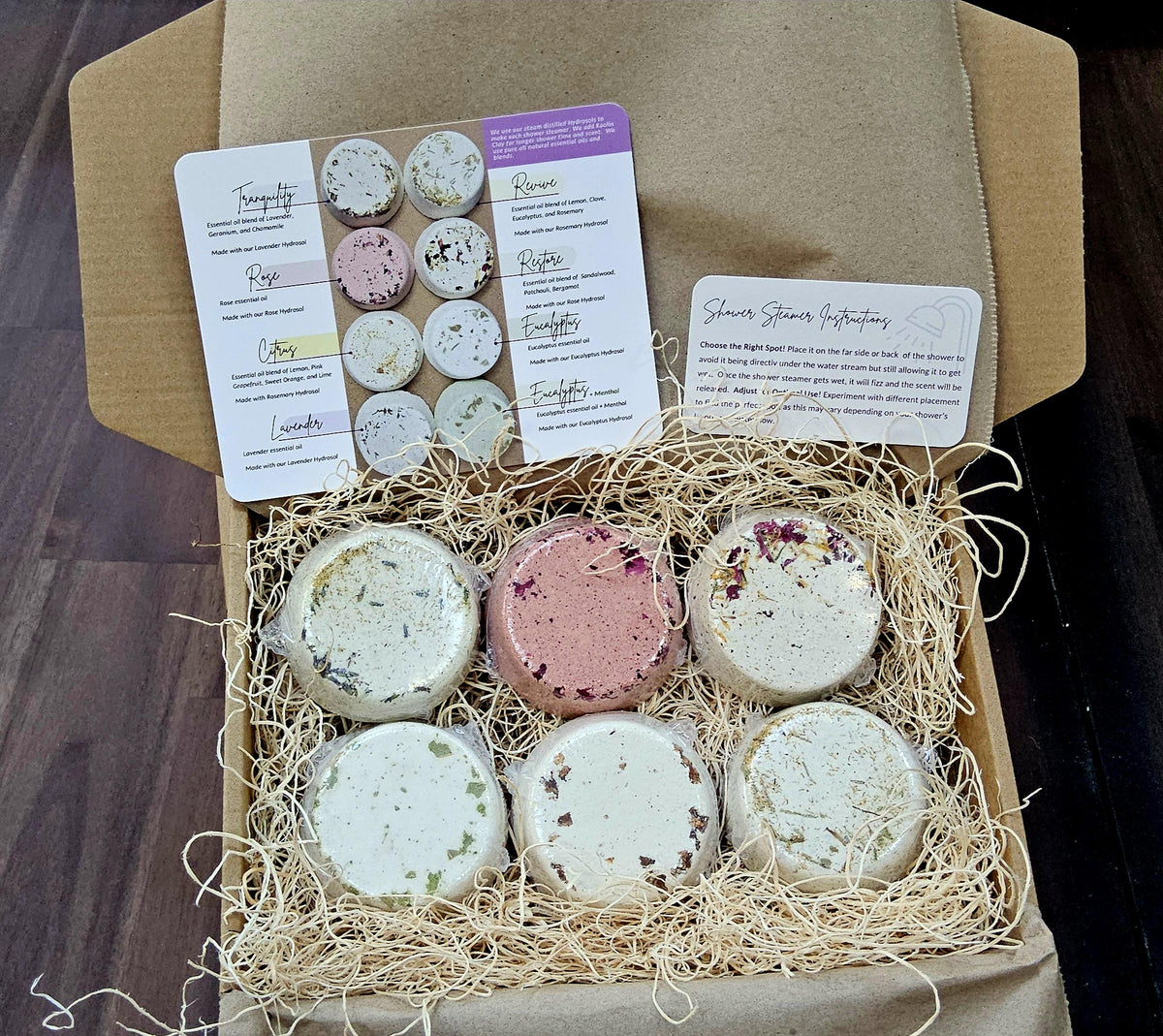 InspireByMK shower steamer gift set featuring six handcrafted aromatherapy steamers in kraft paper box with natural straw packing, product information cards listing scents including Tranquility, Rose, Citrus, Lavender, Revive, Restore, and Eucalyptus with essential oils and hydrosols, shower steamer instructions included