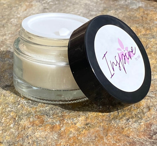 InspireByMK balm product featuring open clear glass jar with black lid showing creamy pale balm texture and purple Inspire label, handcrafted natural skincare moisturizer displayed on stone surface