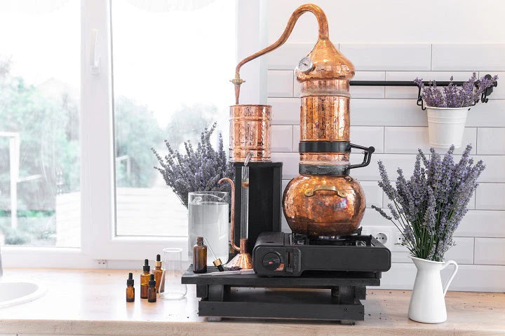 Copper distillation still setup for making pure hydrosols, featuring a large handcrafted copper alembic still with condenser on a black base, surrounded by fresh lavender bunches, small amber dropper bottles, glass jar and white ceramic pitcher on a bright kitchen counter. Authentic small-batch steam distillation process behind our all natural organic facial mist, body spray and aromatherapy products.