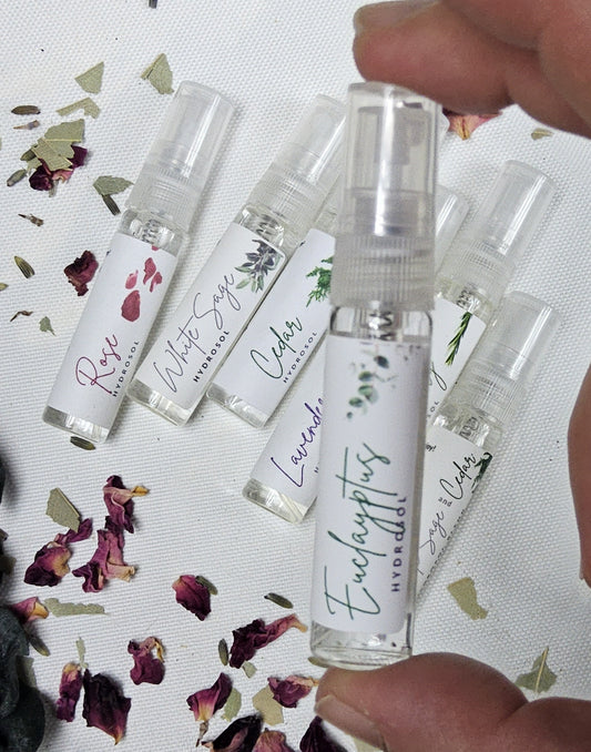 Hand holding multiple trial size hydrosol spray bottles including Eucalyptus, White Sage, Cedar, and more. Small sample size all-natural organic hydrosols perfect for travel, gifting, or trying different scents.
