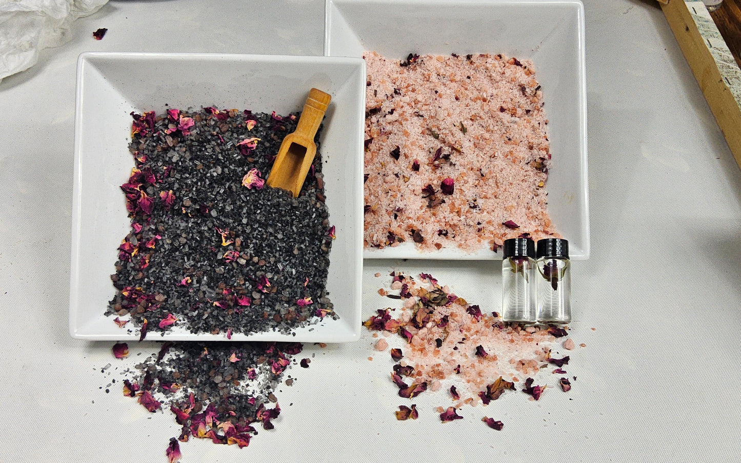 InspireByMK Rose bath salts displayed in two square white dishes — the Lava Blend on the left with dark mineral-rich salts and a wooden scoop, and the White Blend on the right with pink Himalayan and Epsom salts, both topped with dried rose petals alongside two complimentary bath oil bottles