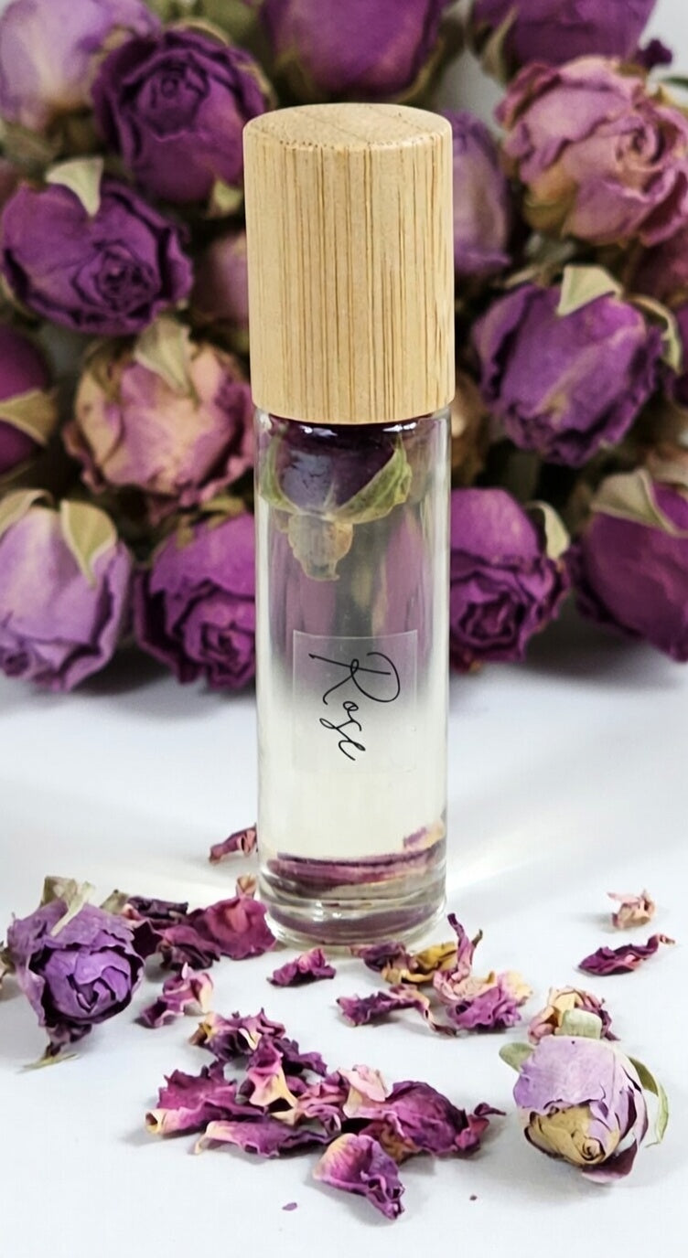 Elegant Rose essential oil roller bottle with real dried rose buds inside, set against a backdrop of lush purple roses and scattered petals. Handcrafted all-natural aromatherapy roller made with pure rose essential oil, vitamin E, and sweet almond oil for romantic and calming vibes.