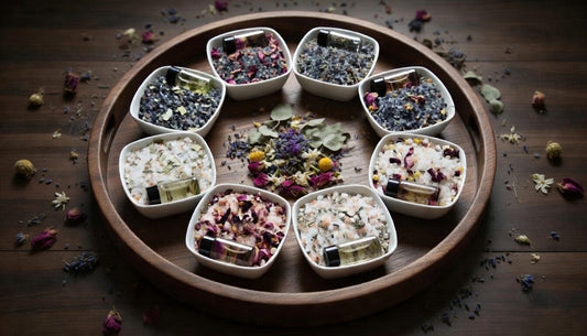 InspireByMK handcrafted bath salts collection featuring eight varieties in white ceramic bowls with dried rose petals, lavender, chamomile, and botanicals, gold essential oil roller bottles, arranged on wooden display tray for natural aromatherapy and spa wellness