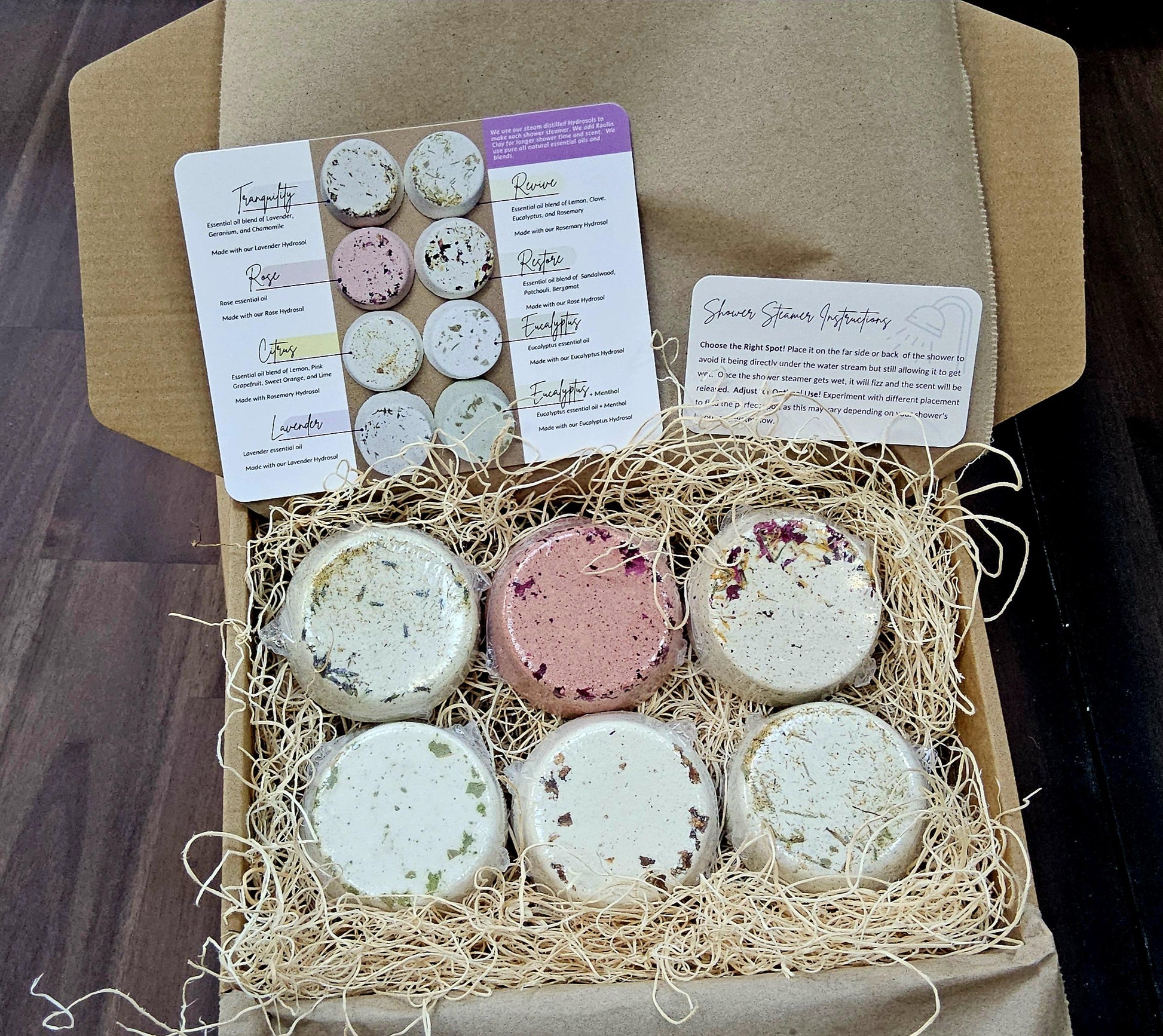 InspireByMK shower steamer gift set featuring six handcrafted aromatherapy steamers in kraft paper box with natural straw packing, product information cards listing scents including Tranquility, Rose, Citrus, Lavender, Revive, Restore, and Eucalyptus with essential oils and hydrosols, shower steamer instructions included