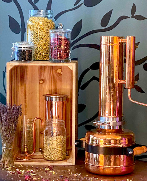 Traditional copper distillation still called "Betsy" used to steam distill our pure hydrosols, shown with jars of dried lavender, rose petals, and other botanicals. Handcrafted all-natural hydrosol production process for authentic aromatherapy products.