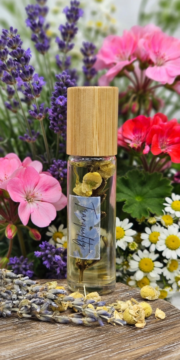 Tranquility essential oil roller bottle surrounded by fresh chamomile flowers, lavender, and geranium blooms. Calming all-natural aromatherapy roller handcrafted with lavender, geranium, and roman chamomile essential oils plus dried botanicals in vitamin E and sweet almond oil base for peaceful relaxation.